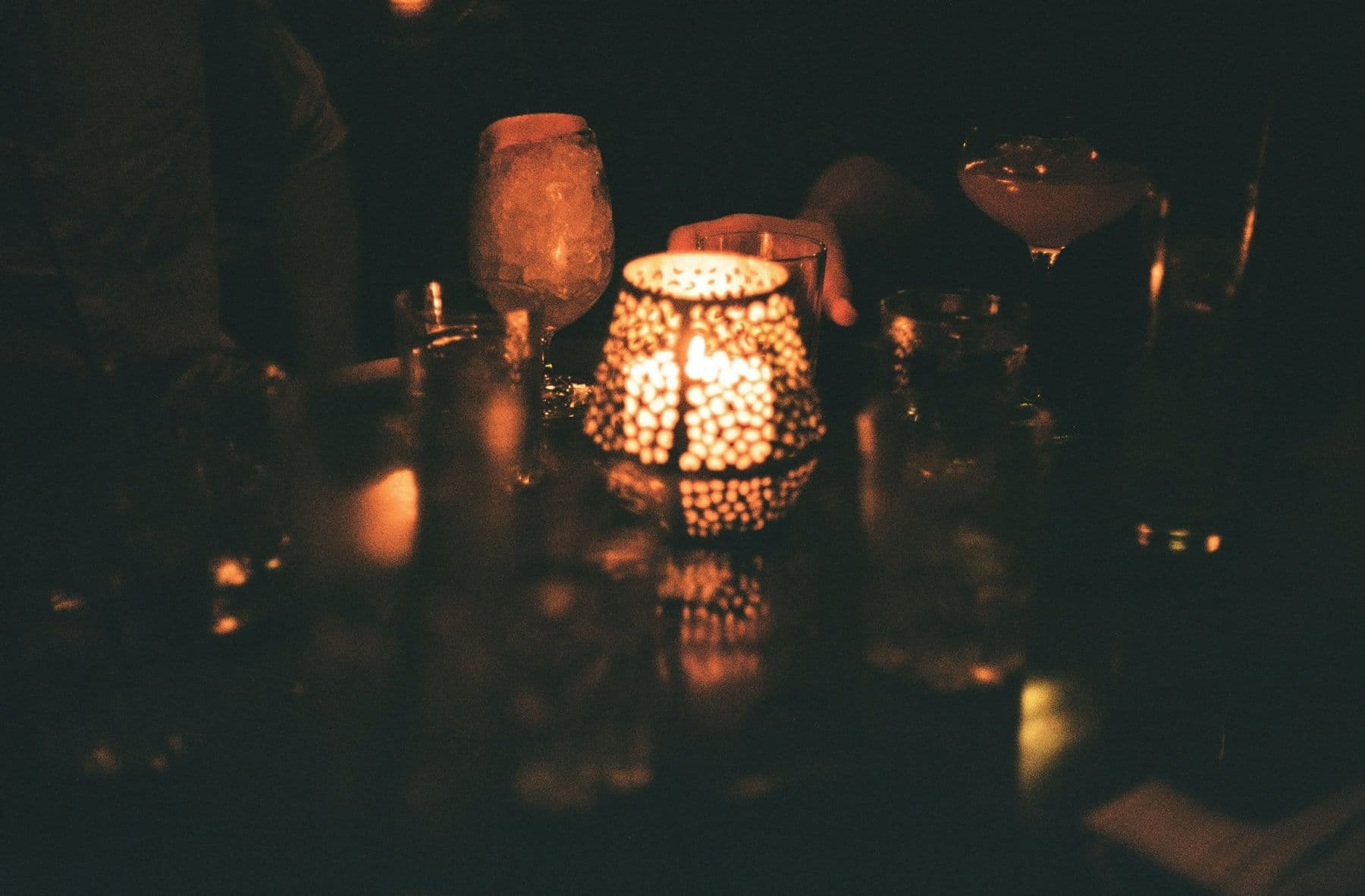 A candlelit table in a warm bar, glasses gathered close, a hand reaching in from the edge of frame